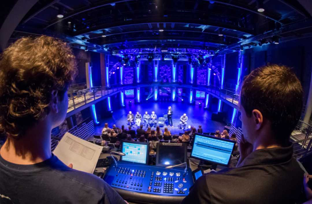 Over-the-shoulder image of two men at a light board overlooking a stage with blue lights