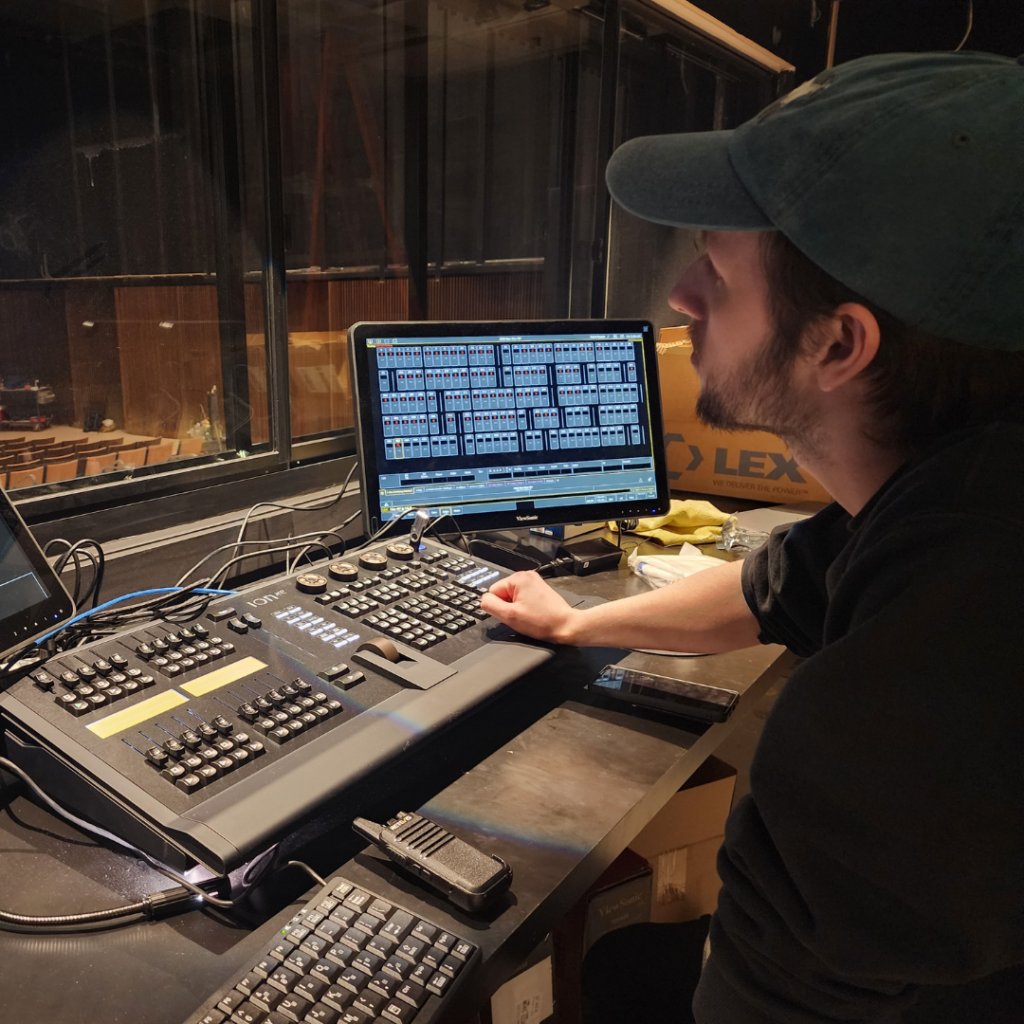 A man wearing a black shirt and a grey hat sitting at a light board.